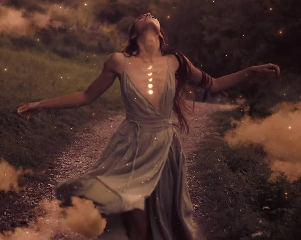 Enchanting woman with arms outstretched, wearing a flowing dress and standing on a misty path, illuminated by glowing light orbs. Her head is tilted back in a pose of surrender and freedom, evoking themes of spiritual awakening, feminine empowerment, and connection with the divine. The ethereal scene captures a sense of magic, transformation, and the wild feminine spirit.