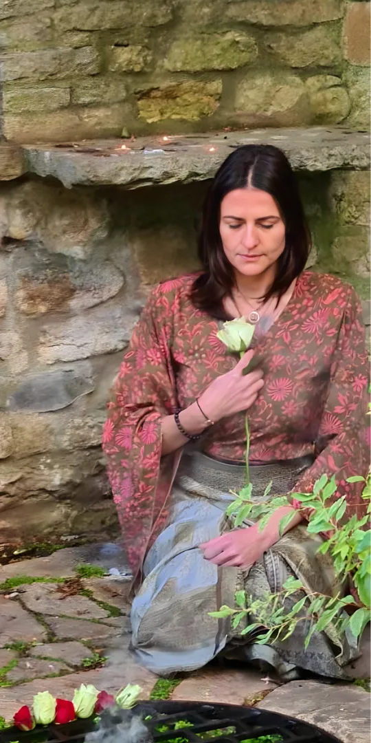 Woman seated in meditation, holding a white rose close to her heart, wearing a patterned floral shawl. The scene takes place in a sacred space with a stone wall backdrop, surrounded by elements such as rose petals, greenery, and smoldering incense. The image evokes themes of feminine healing, sacred rituals, and deep spiritual connection. It reflects the journey of self-exploration, heart-centered practices, and embracing the divine feminine, aligned with sacred embodiment and spiritual awakening.