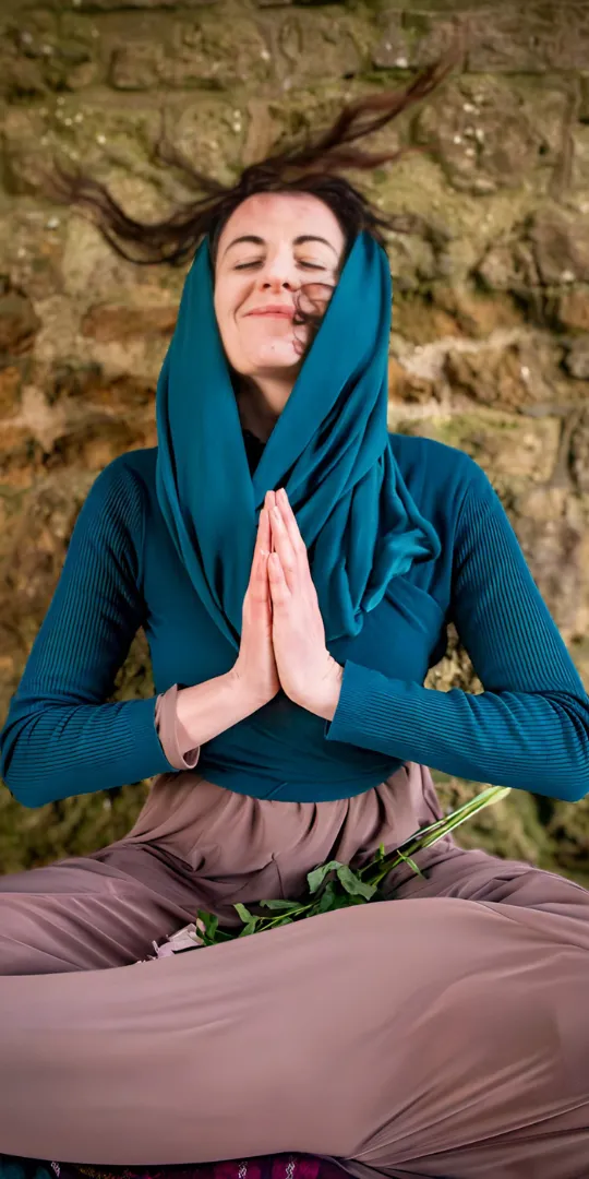 Woman sitting in a meditative pose with hands in prayer position, wearing a teal shawl draped over her head. Her eyes are closed with a serene expression, symbolizing inner peace, spiritual awakening, and sacred feminine embodiment. A single rose rests across her lap, evoking themes of heart-centered healing, deep self-exploration, and connection to the divine feminine. The stone wall backdrop adds a grounding element, highlighting the blend of ancient wisdom and modern spiritual practices.