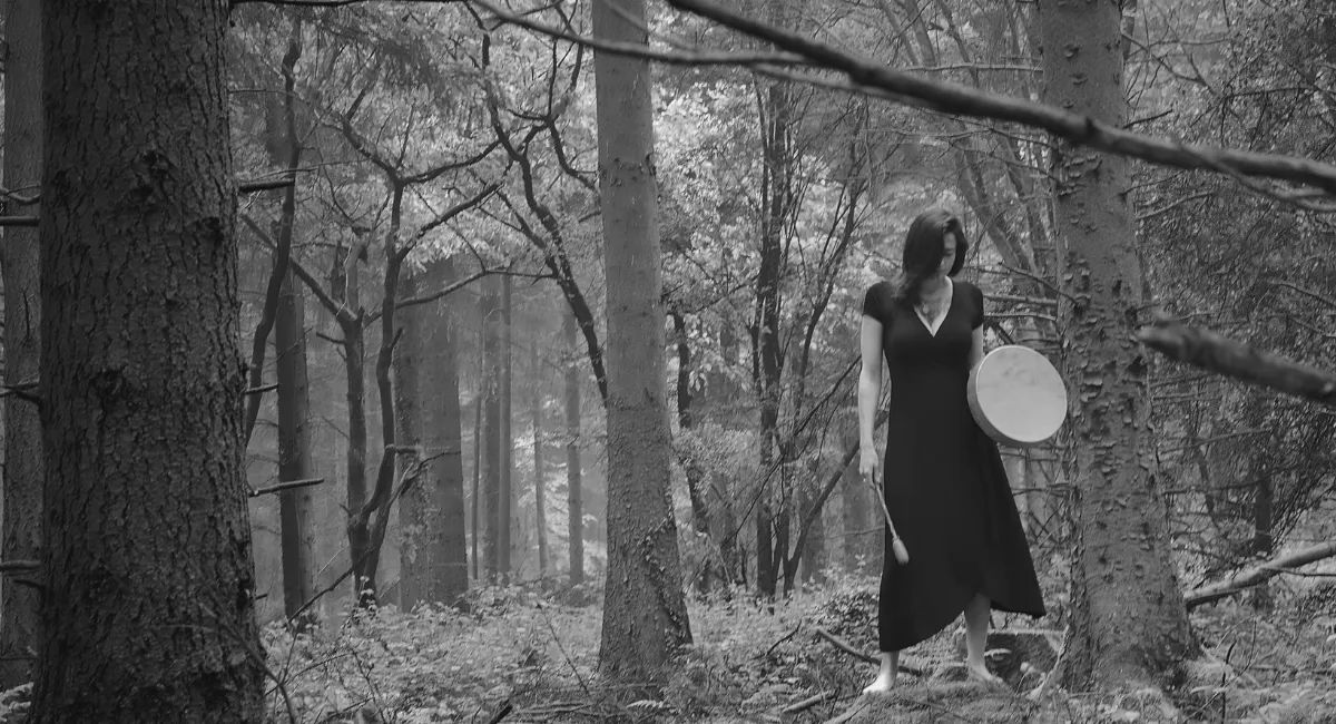 Woman in forest drumming for deep feminine exploration, connecting with nature’s wisdom and embodying sacred healing rituals.