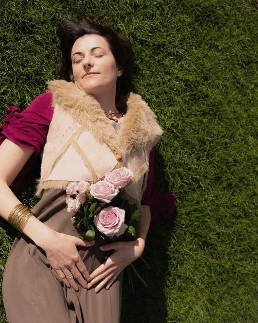 Woman lying on grass, holding pink roses over her womb, symbolizing feminine healing, sacred embodiment, and deep connection to divine love.