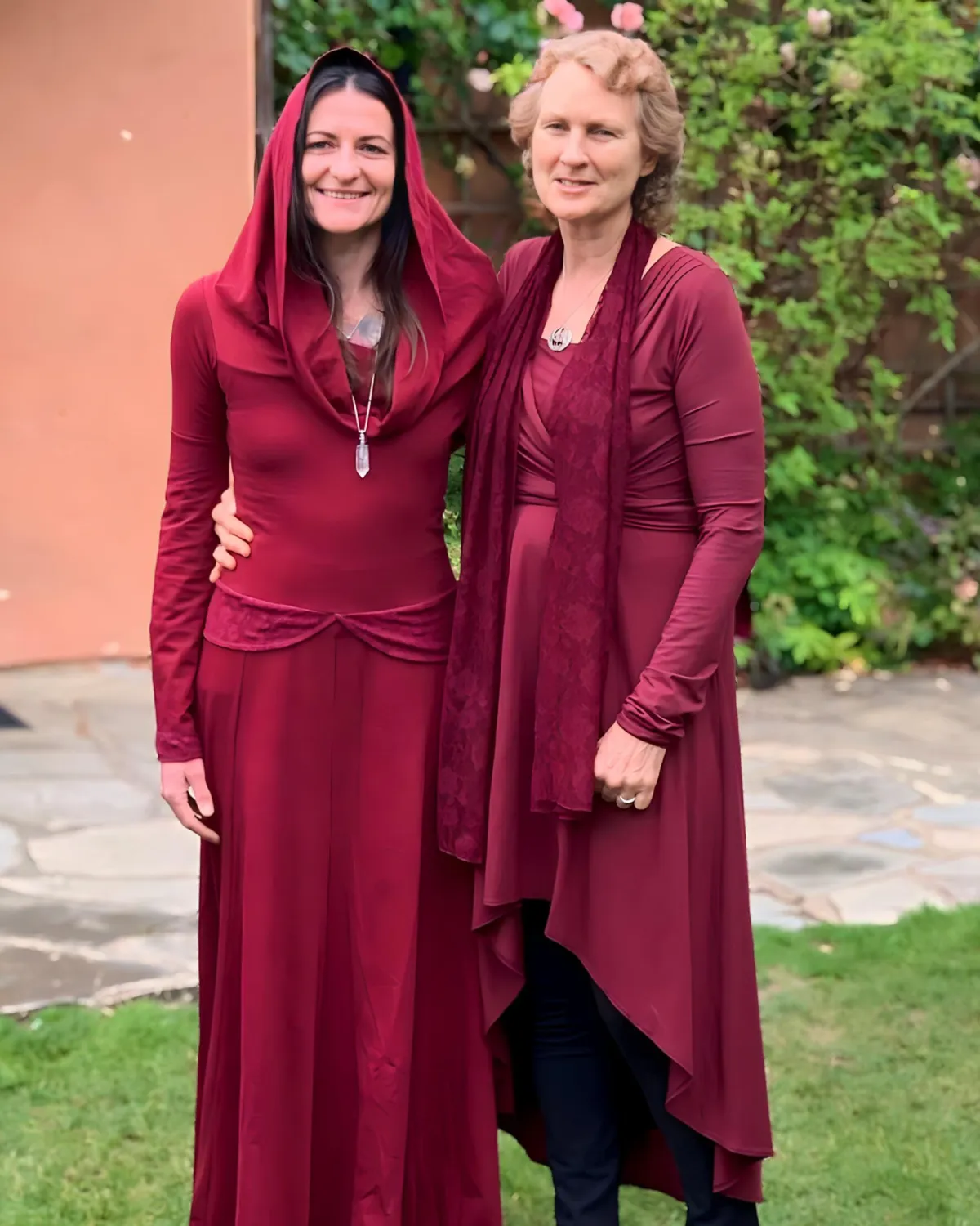 Two women in red gowns, symbolising sisterhood, sacred feminine connection, and shared spiritual journey, standing in a garden setting with greenery.