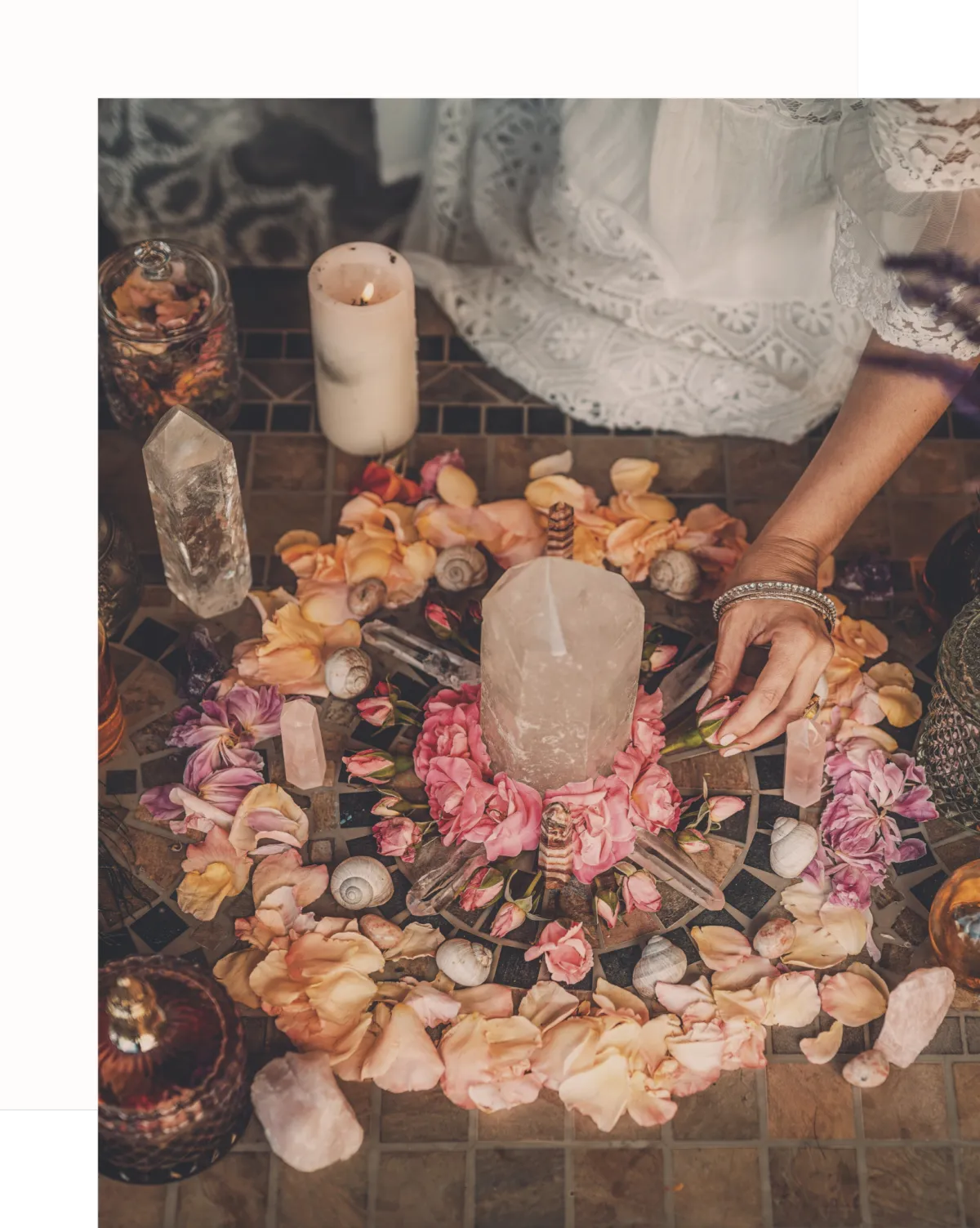 Sacred altar arrangement with crystals, flowers, rose petals, seashells, and candles, used for spiritual rituals and healing ceremonies. A woman’s hand reaches toward the altar, wearing a white lace dress, evoking themes of feminine energy, sacred space, and intentional ritual. The scene suggests a connection to earth-based spirituality, crystal healing, and the divine feminine, creating a sense of beauty and reverence.