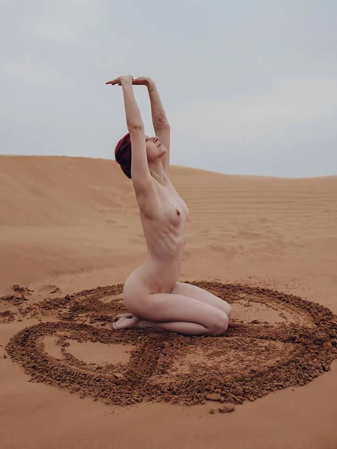 Nude woman kneeling in a desert, surrounded by a sacred earth symbol, embodying feminine freedom, spiritual awakening, and deep-rooted connection to nature.