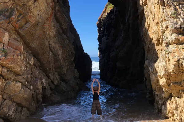 Woman standing in a powerful pose with arms raised, facing a narrow passage between two towering rock cliffs by the ocean. The scene captures the essence of feminine strength, spiritual awakening, and connection to nature. The image evokes themes of transformation, sacred embodiment, and the journey of returning to one’s inner power, resonating with the path of deep self-exploration and embracing the wild, untamed feminine. The coastal setting adds an element of grounding and natural beauty, symbolizing the merging of earth and spirit.