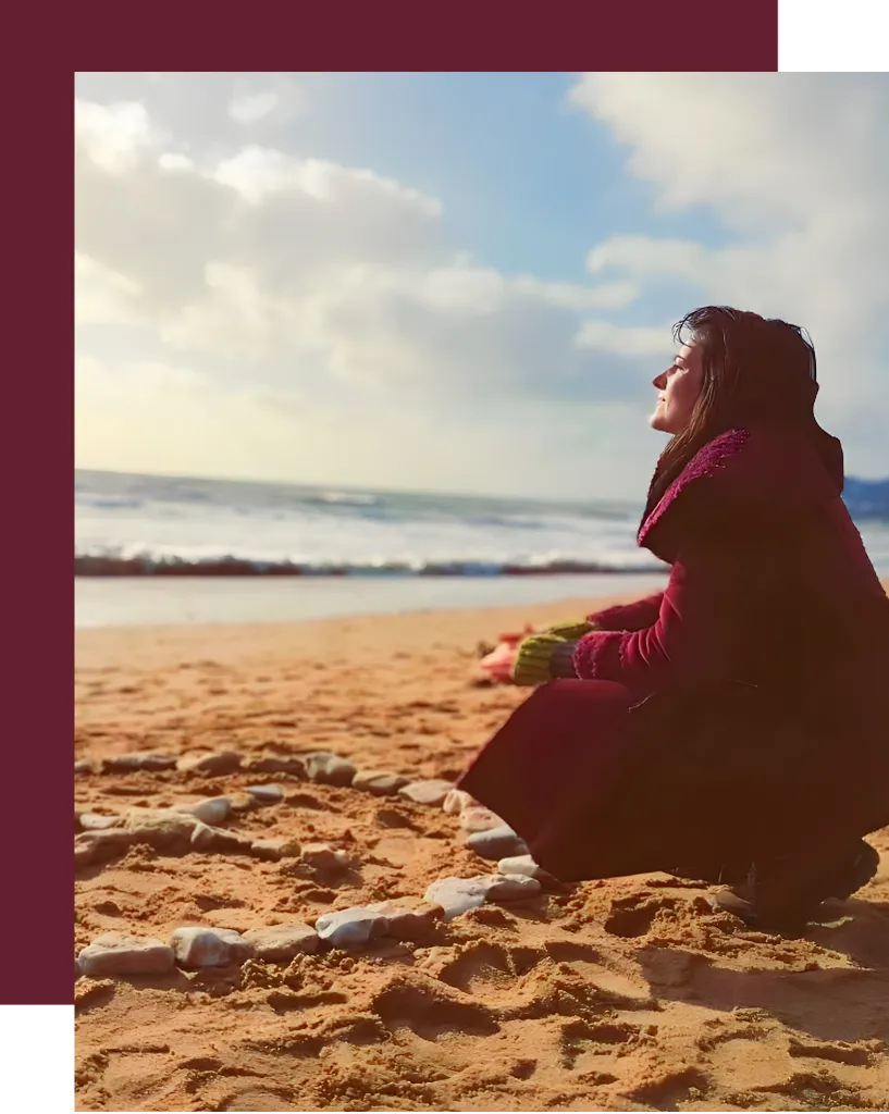 Woman kneeling on a sandy beach near a circle of stones, symbolizing grounding, sacred rituals, and connecting with nature and the divine feminine.