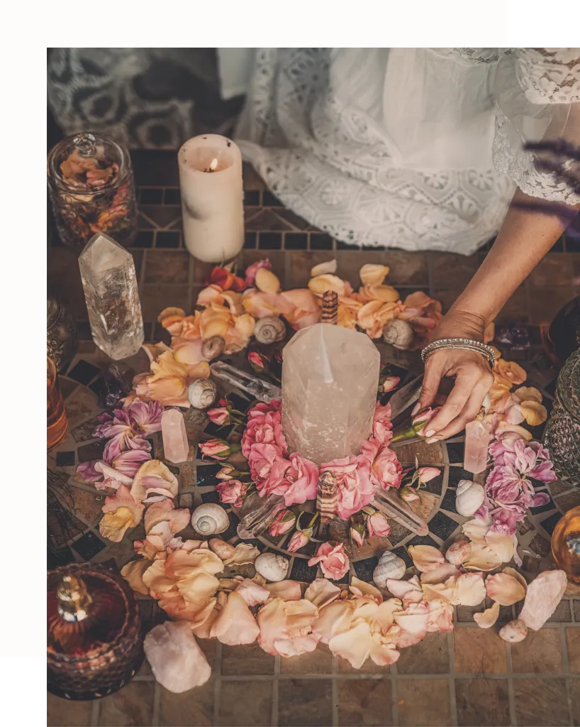 Sacred altar arrangement with crystals, flowers, rose petals, seashells, and candles, used for spiritual rituals and healing ceremonies. A woman’s hand reaches toward the altar, wearing a white lace dress, evoking themes of feminine energy, sacred space, and intentional ritual. The scene suggests a connection to earth-based spirituality, crystal healing, and the divine feminine, creating a sense of beauty and reverence.