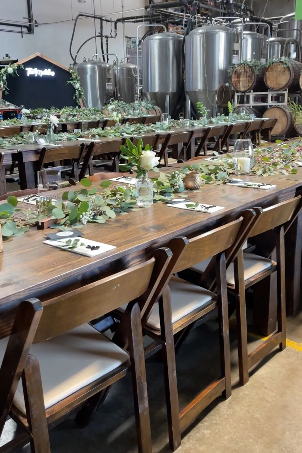 Long wooden tables with greenery runners set for a party next to the brewing tanks