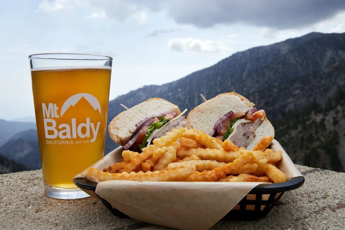A pint of Mt Baldy beer next to a basket of loaded fries and a sandwich, with mountains behind