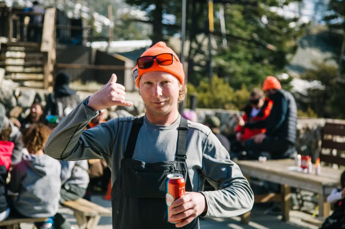 A smiling guest in an orange beanie gives a thumbs up with a beer on the mountain
