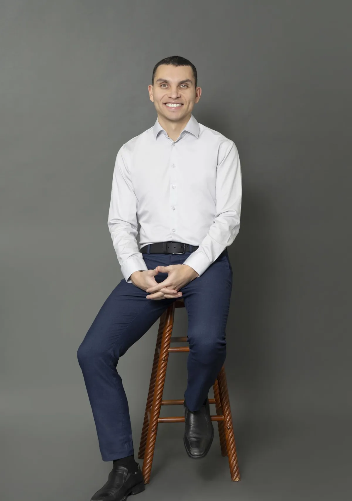 Chris Stamler seated on a wooden stool, smiling with a calm, approachable expression, wearing a light button-up shirt and dark dress pants.