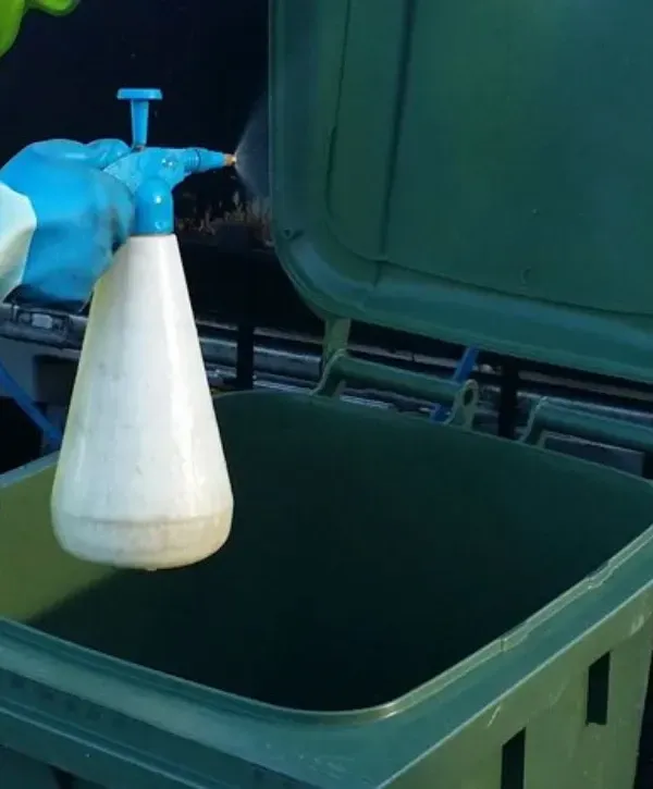 Worker spraying disinfectant inside a green trash bin