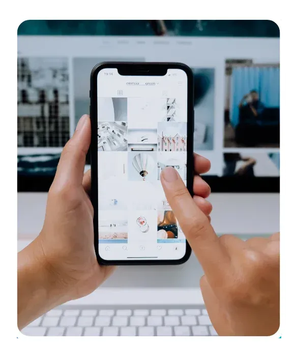 Person managing social media posts on a smartphone with visual content displayed on a desktop screen.