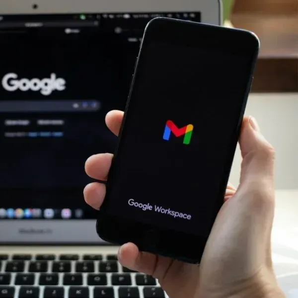 Person holding a smartphone displaying the Google Workspace Gmail app with Google search open on a laptop in the background.
