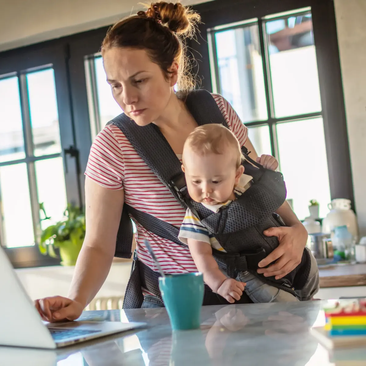 Mom with baby in carrier checking meal plan on laptop in a bright kitchen, symbolizing real-life dinner prep for busy moms..