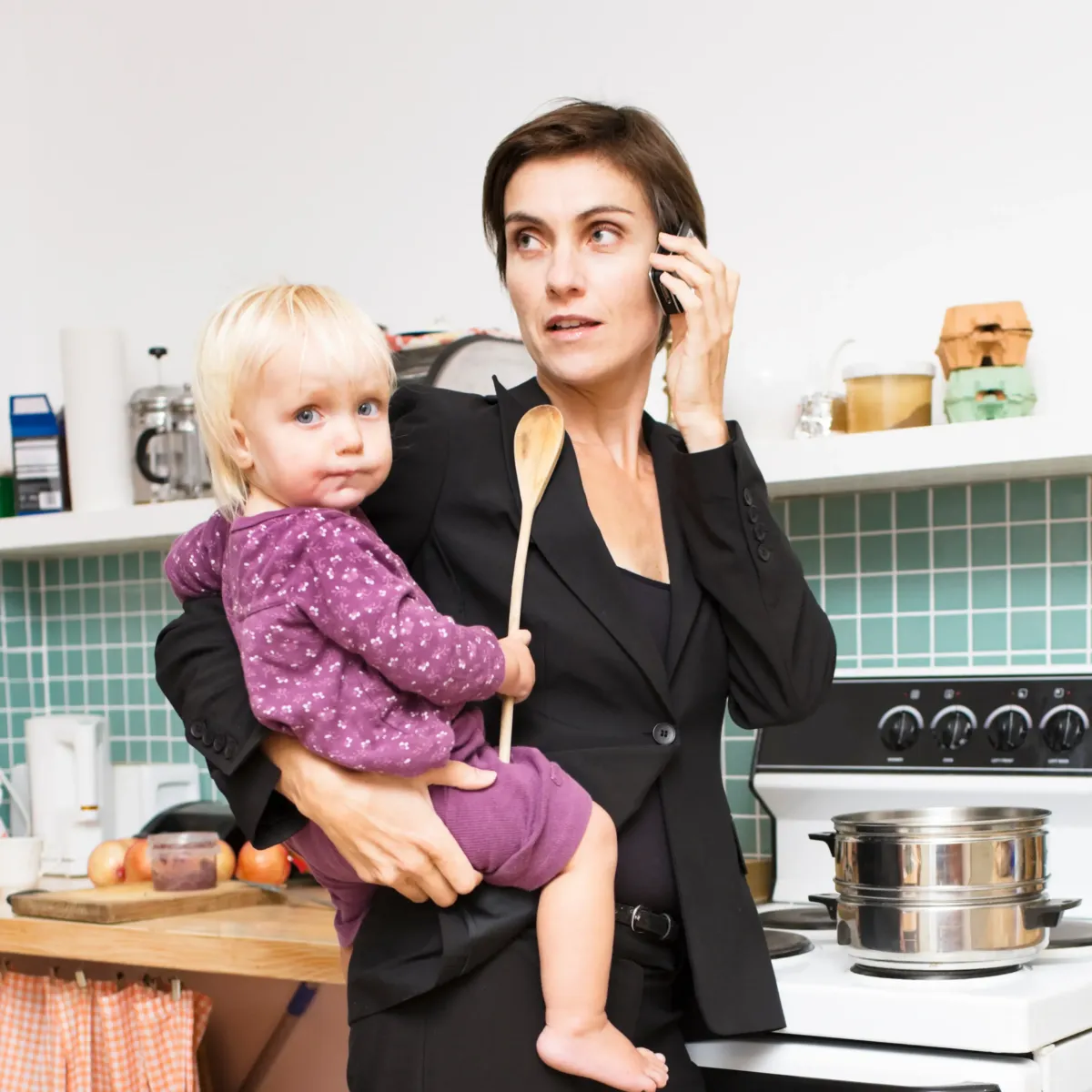 Busy mom in the kitchen holding a toddler and talking on the phone, representing real-life meal planning for overwhelmed mothers.