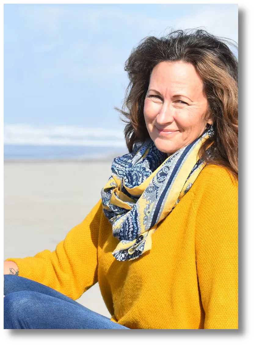 Portrait photograph of a woman sitting on a beach with the ocean behind her, wearing a bright yellow sweater and a blue-and-yellow patterned scarf.