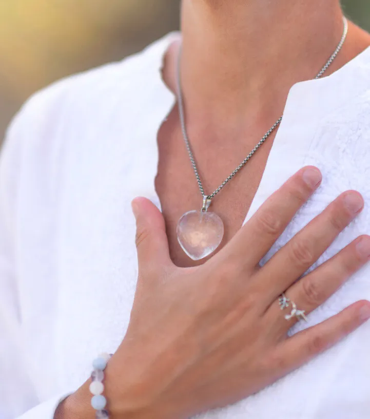 Close-up photograph of a person wearing a white blouse, with their hand resting over their heart, wearing a clear heart-shaped pendant necklace.