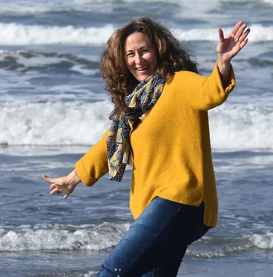 Portrait of a woman with curly brown hair wearing a bright yellow sweater and blue jeans, standing outdoors near a rocky beach with her arms outstretched cheerfully