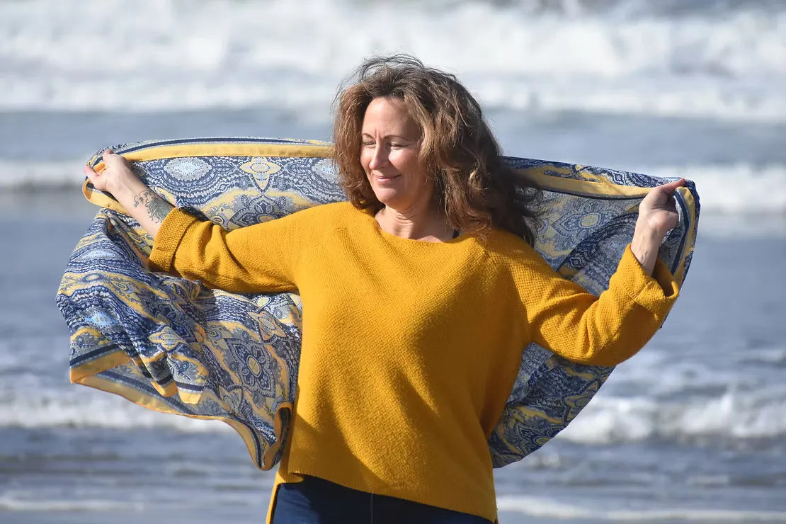 A smiling woman with brown hair standing near a beach, wearing a mustard yellow sweater, holding a blue and yellow patterned scarf open behind her catching the wind, with waves visible in the background
