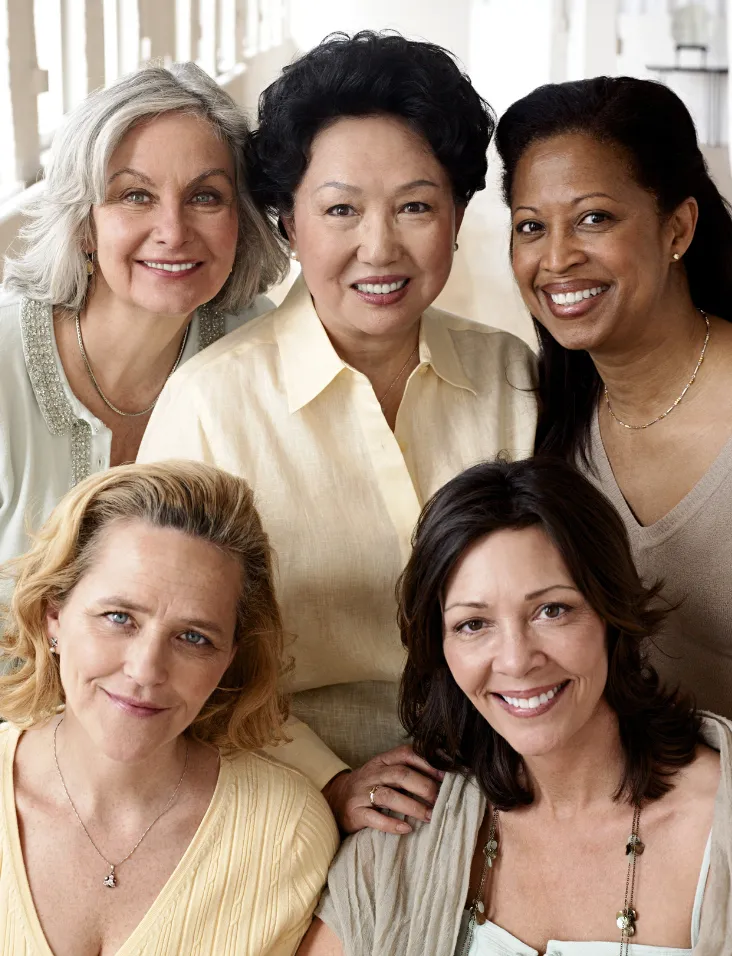 Portrait of five women of varying ages smiling together in a warm, supportive group pose; soft daylight, neutral clothing, friendly and welcoming mood.