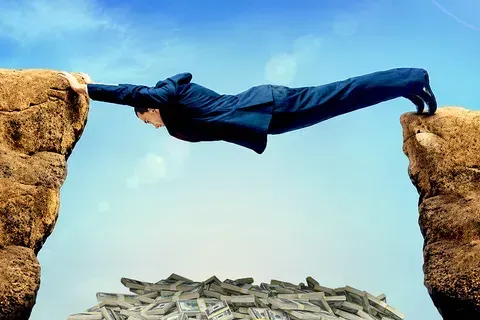 Businessman stretches across a rock gap above a pile of money under a clear blue sky.