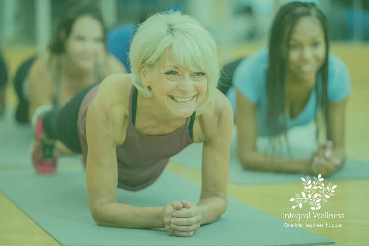 A group of adults in a bright fitness studio holding a plank position on yoga mats. One person is in focus at the front while others line up behind them. The atmosphere looks supportive and energetic. The Integral Wellness logo appears in the bottom right corner with the tagline “One life: healthier, happier.”