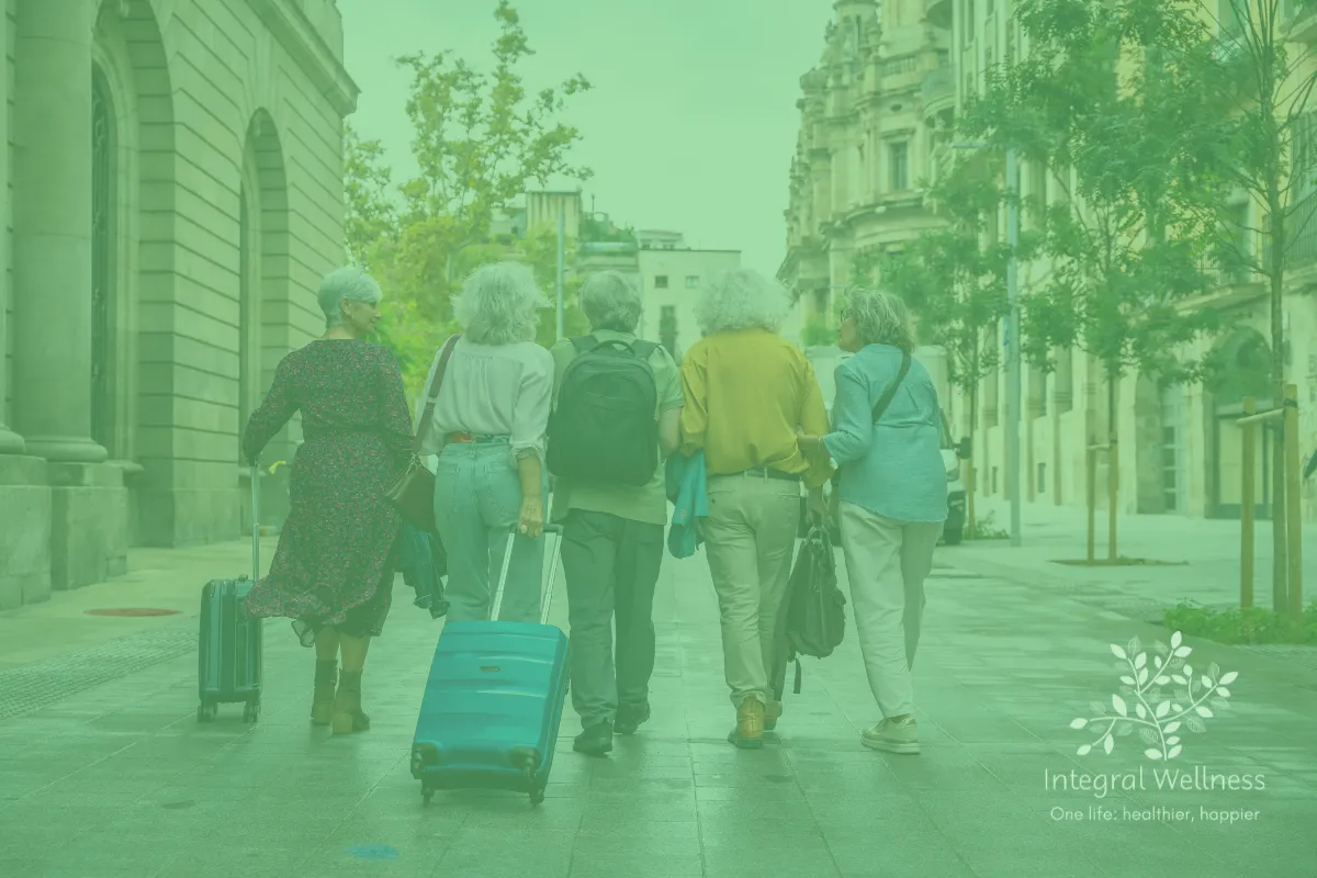 Five older adults walking together down a city street with luggage, surrounded by tall buildings and trees. The image has a green overlay and includes the Integral Wellness logo with the tagline 'One life: healthier, happier' in the corner