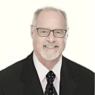 A friendly male financial advisor in his 40s, smiling and answering a phone call at a bright office desk with a notepad and pen, cityscape visible through the window. 3:2 aspect ratio, professional, approachable.