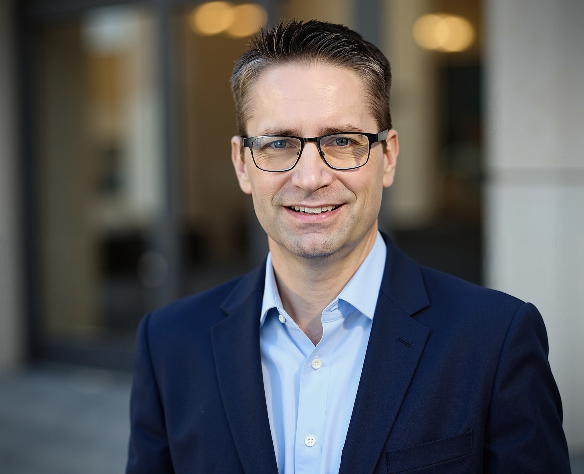 A friendly male financial advisor in his 40s, smiling and answering a phone call at a bright office desk with a notepad and pen, cityscape visible through the window. 3:2 aspect ratio, professional, approachable.