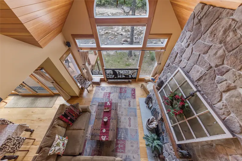 Overhead view of a vaulted living room with a stone fireplace wall, wood ceiling, tall windows and a rustic sofa
