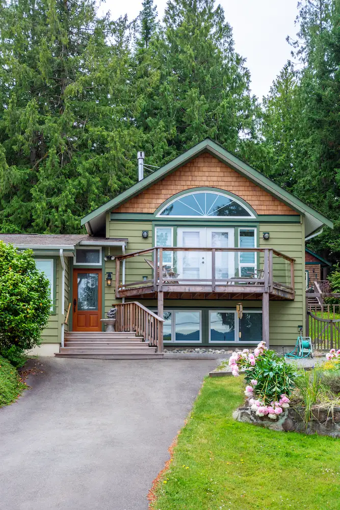 Green two-storey house with cedar shake gable, arched window, wooden deck and front stairs surrounded by evergreen trees