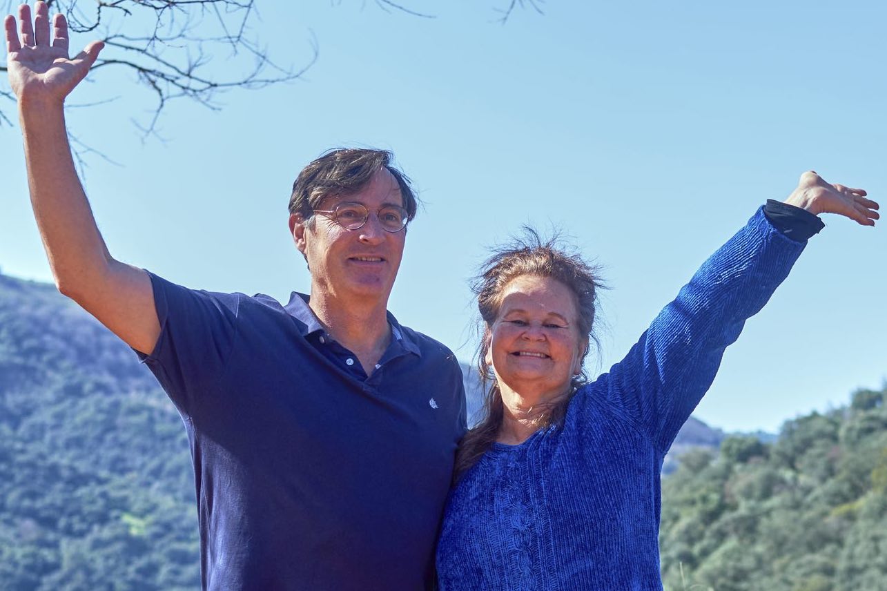 Two experienced retreat leaders, one male and one female, standing together in a cozy lodge interior with large windows, warm wooden accents, and soft natural light, both smiling and exuding a welcoming, supportive presence.