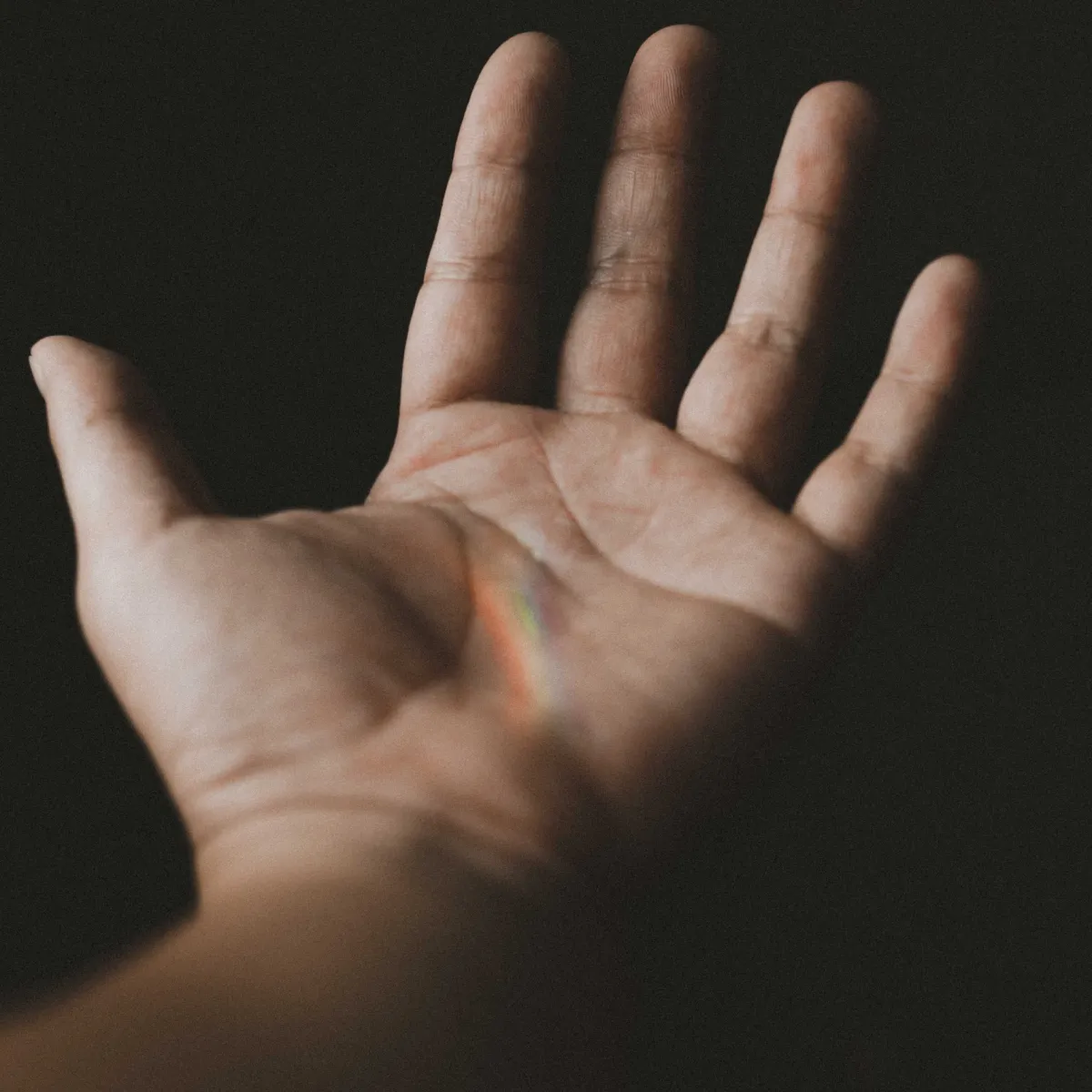 A close-up of an open hand with a faint rainbow light across the palm. The dark background highlights the hand and the soft glow of the rainbow, symbolizing hope, spirituality, and connection. The image evokes a sense of introspection and inner energy.