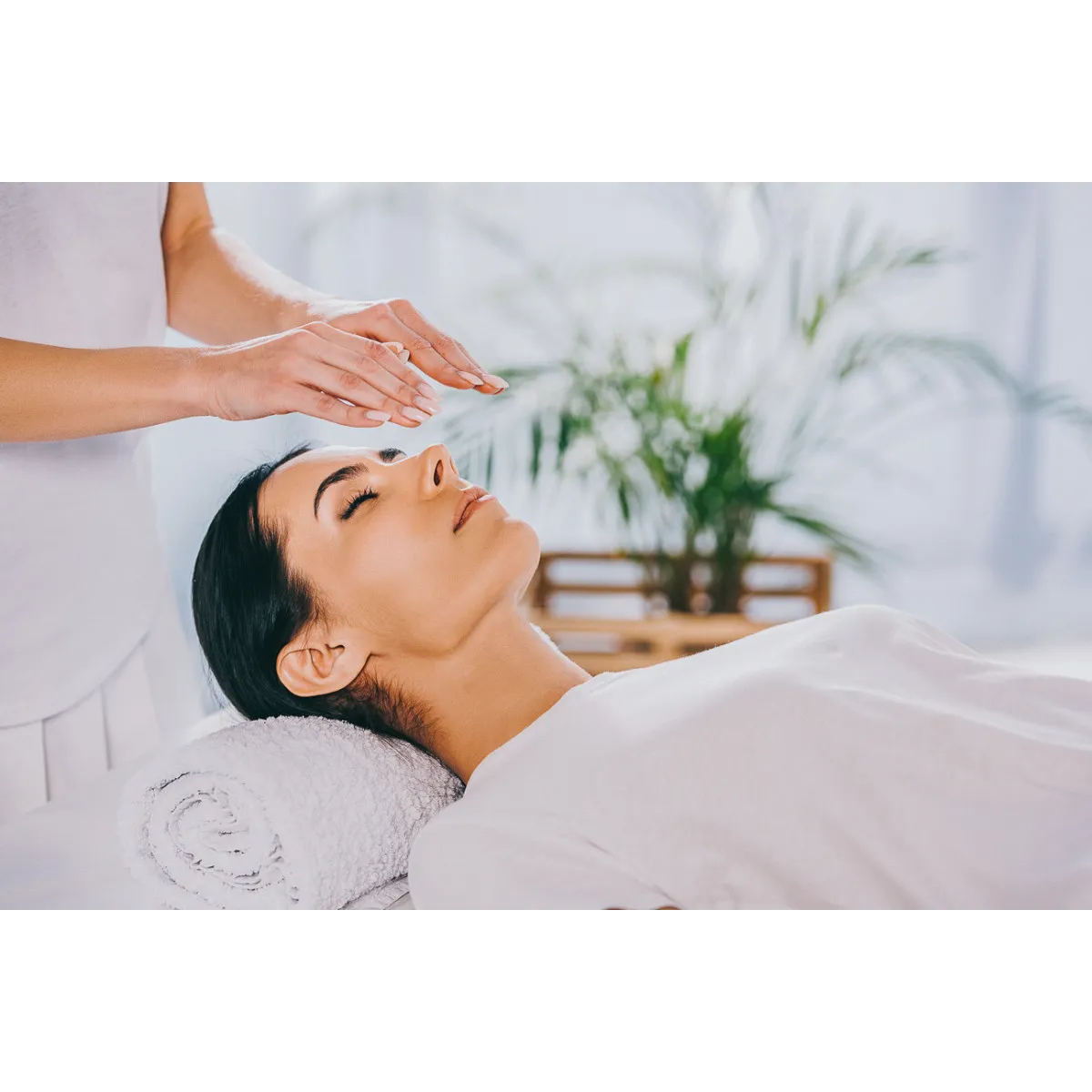 A serene scene of a woman receiving energy healing or reiki. She is lying down with her eyes closed, while a healer hovers their hands above her, channeling healing energy. The environment is peaceful, with soft lighting and a plant in the background, emphasizing relaxation and spiritual balance.