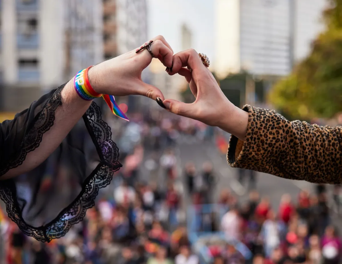 Two hands forming a heart shape in the foreground, symbolizing love and unity. One hand wears a rainbow-colored pride wristband, and the scene takes place in a public event or pride parade, with a blurred crowd in the background. The image celebrates LGBTQ+ love, pride, and solidarity.