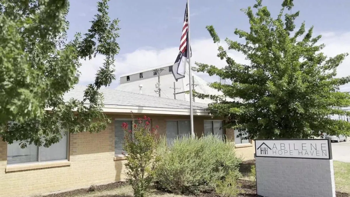 Front view of Abilene Hope Haven’s building with a U.S. flag and Christian flag flying on a pole, surrounded by green trees and shrubs, with a white sign displaying the organization's name near the entrance