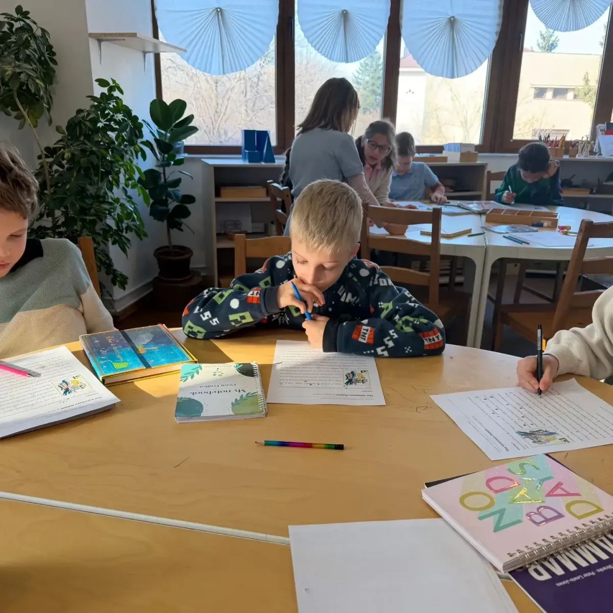 Profesor care îi îndrumă pe doi copii de vârstă școlară primară folosind materiale Montessori la o masă joasă.