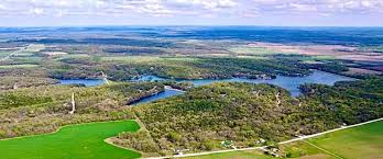 An aerial view of a wide landscape with rolling fields, forests, and a large lake in the distance.