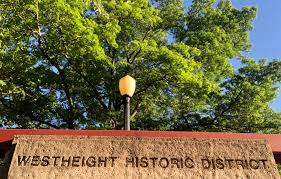 A sign reading ‘Westheight Historic District’ mounted on a brick wall beneath green leafy trees.