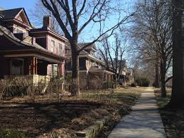 A neighborhood sidewalk lined with older homes and tall leafless trees on a clear day.