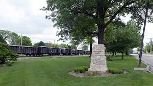 A stone monument in a grassy park with trees and a train passing in the background