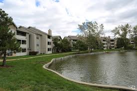 An apartment community with multi-story buildings surrounding a pond with a walking path and trees.