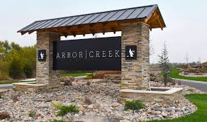 A stone entrance sign reading ‘Arbor Creek’ beneath a wooden roof structure surrounded by landscaping and gravel