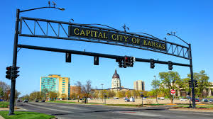 A large arched sign reading ‘Capital City of Kansas’ above a roadway, with city buildings in the background.