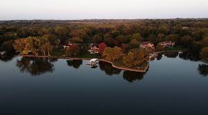 An aerial view of a calm lake surrounded by trees with autumn foliage and scattered homes.
