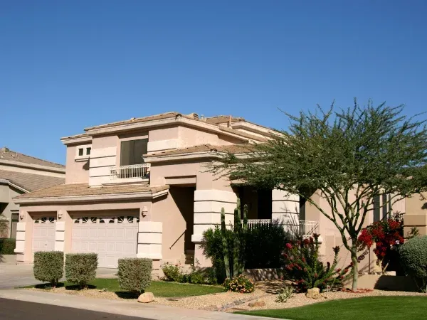 A two-story desert home facade with light stucco, a double garage, and xeriscaped landscaping under a clear blue sky.