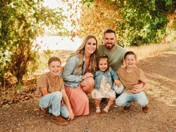 A happy family of five, including parents and three young children, poses smiling on a sunlit autumn path outdoors.