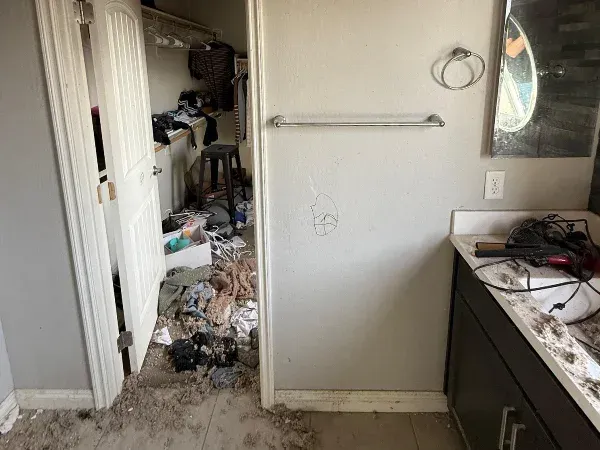A distressed bedroom closet, full of clothes and debris, next to a messy bathroom sink area with a dirty counter.