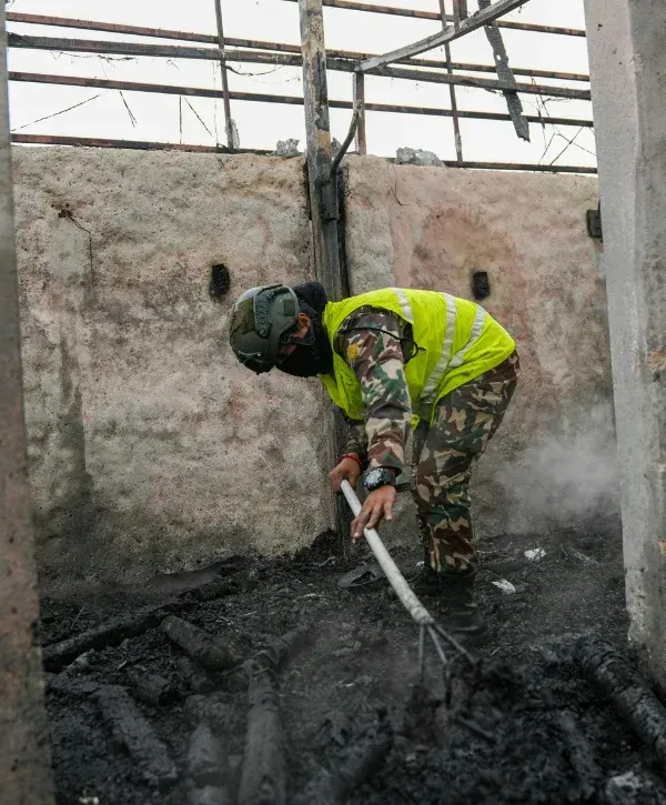 A person in tactical gear and a high-vis vest raking charred debris on a smoky, damaged concrete floor.